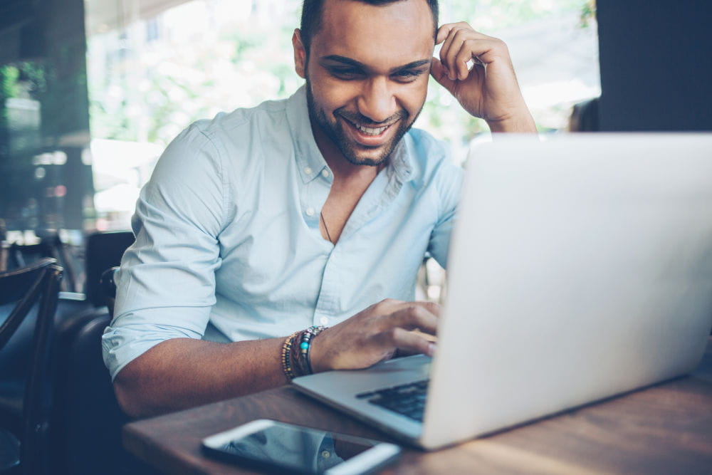 Un employé assis à une table, souriant tout en utilisant un ordinateur, affichant une attitude positive dans un environnement de travail.